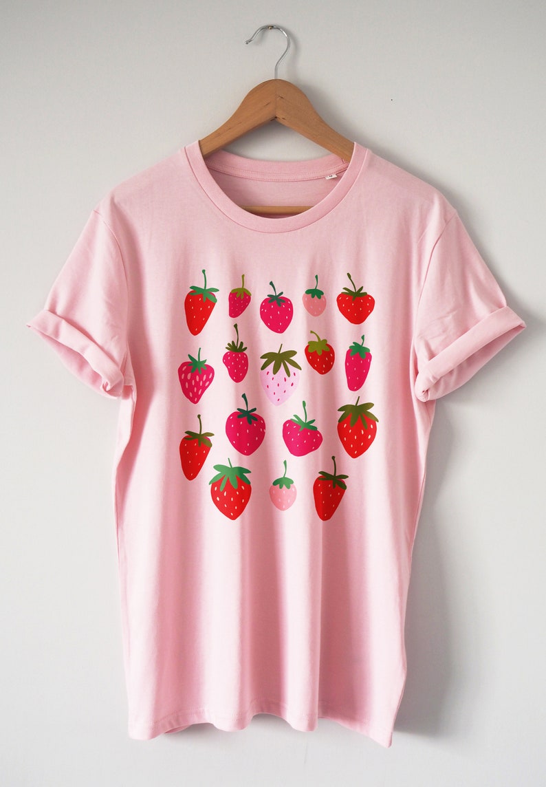 Puede incluir: Una camiseta rosa con un estampado gr&aacute;fico de fresas rojas, rosas y verdes.