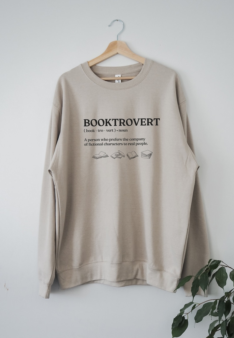 May include: A beige sweatshirt with the text "BOOKTROVERT (book-tro-vert) noun A person who prefers the company of fictional characters to real people." and a graphic of several open books.