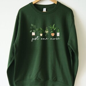 Puede incluir: Una sudadera verde oscuro con un gr&aacute;fico blanco de plantas en macetas y el texto "just one more".