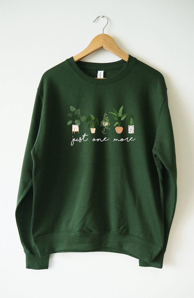 Puede incluir: Una sudadera verde oscuro con un gr&aacute;fico blanco de plantas en macetas y el texto "just one more".