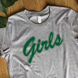 May include: Grey t-shirt with a green "Girls" graphic in a retro font.