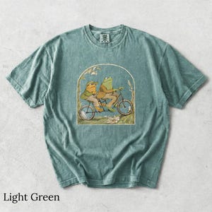 May include: A light green short-sleeve t-shirt featuring a graphic illustration of two frogs riding a tandem bicycle. The frogs are wearing brown jackets and hats. The design is framed by an archway with floral accents at the base.