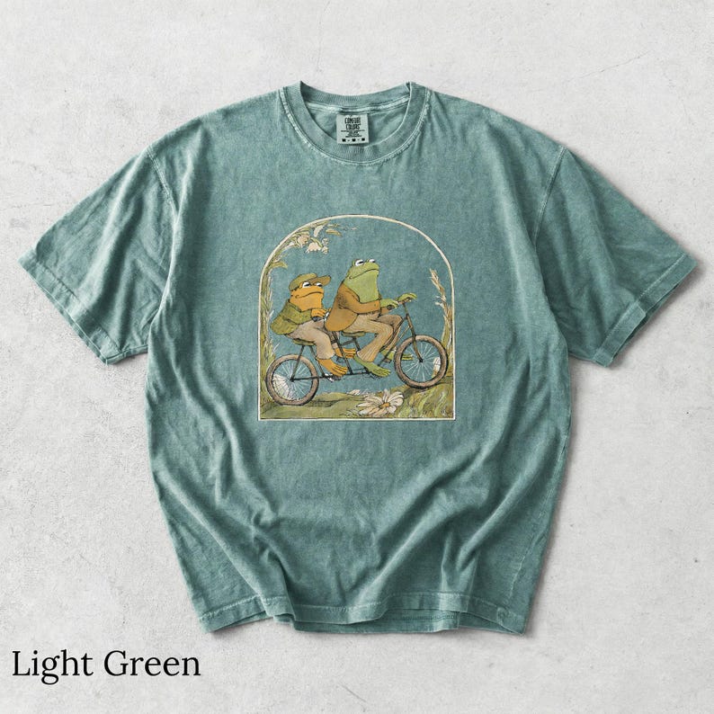 May include: A light green short-sleeve t-shirt featuring a graphic illustration of two frogs riding a tandem bicycle. The frogs are wearing brown jackets and hats. The design is framed by an archway with floral accents at the base.
