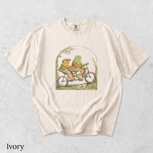 May include: An ivory short-sleeve t-shirt featuring a graphic illustration of two frogs wearing jackets and hats riding a tandem bicycle. The frogs are positioned within an arched frame surrounded by simple line drawings of grass and flowers.