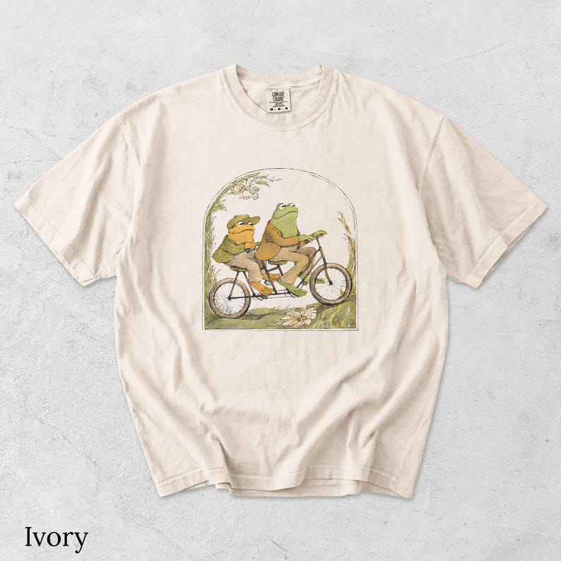 May include: An ivory short-sleeve t-shirt featuring a graphic illustration of two frogs wearing jackets and hats riding a tandem bicycle. The frogs are positioned within an arched frame surrounded by simple line drawings of grass and flowers.