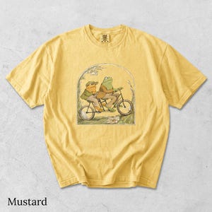 May include: A mustard yellow short-sleeve t-shirt featuring a graphic print of two frogs riding a tandem bicycle. The illustration is framed by an archway with floral details. The shirt has a crew neckline and a Comfort Colors brand label.