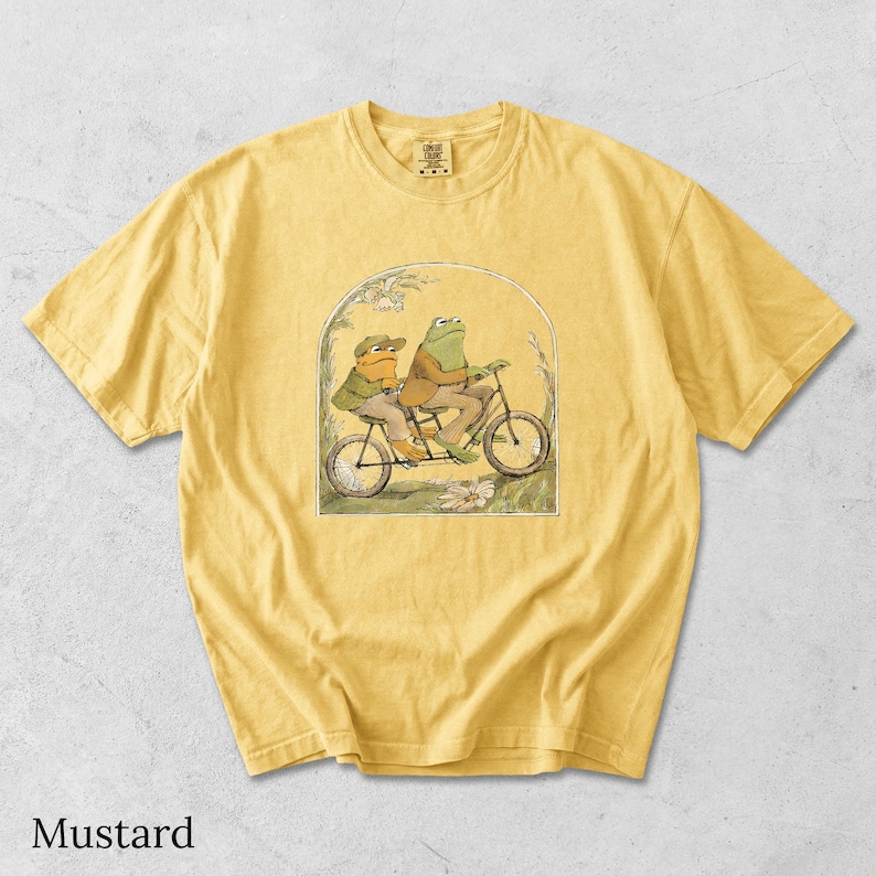 May include: A mustard yellow short-sleeve t-shirt featuring a graphic print of two frogs riding a tandem bicycle. The illustration is framed by an archway with floral details. The shirt has a crew neckline and a Comfort Colors brand label.