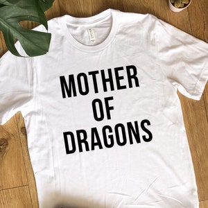 May include: White t-shirt with the text "MOTHER OF DRAGONS" in bold black letters. The shirt has a crew neck and short sleeves. The shirt is made of a soft material.