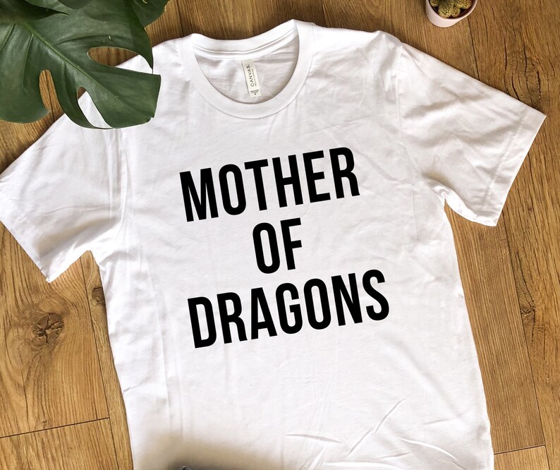 May include: White t-shirt with the text "MOTHER OF DRAGONS" in bold black letters. The shirt has a crew neck and short sleeves. The shirt is made of a soft material.