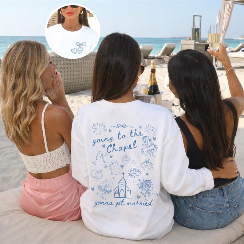 May include: Three people are sitting on a beach. One person is wearing a white sweatshirt with the text "going to the Chapel gonna get married" and a blue illustration. The sweatshirt has a small blue design on the front. The other two people are wearing a white top and a black top.