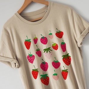 Puede incluir: Una camiseta beige con un estampado gr&aacute;fico de fresas rojas y rosas. Las fresas est&aacute;n dispuestas en un patr&oacute;n aleatorio en la camiseta.
