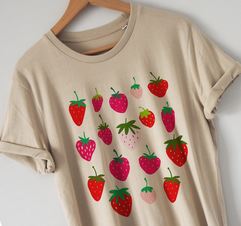Puede incluir: Una camiseta beige con un estampado gr&aacute;fico de fresas rojas y rosas. Las fresas est&aacute;n dispuestas en un patr&oacute;n aleatorio en la camiseta.
