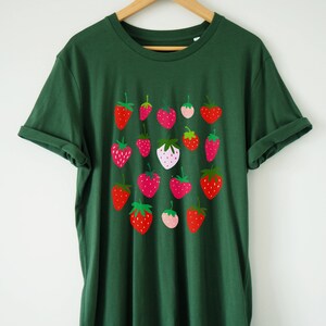 Puede incluir: Una camiseta verde oscuro con un estampado gr&aacute;fico de fresas rojas, rosas y blancas. Las fresas est&aacute;n dispuestas en una cuadr&iacute;cula en la parte delantera de la camiseta.