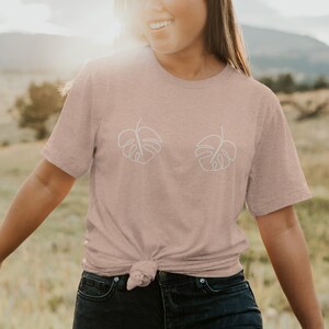 May include: A light pink t-shirt with a white outline of two monstera leaves on the front. The shirt is tied at the waist.
