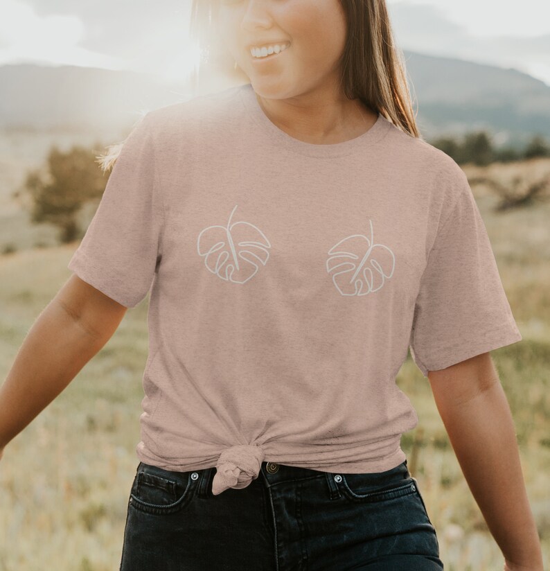 May include: A light pink t-shirt with a white outline of two monstera leaves on the front. The shirt is tied at the waist.