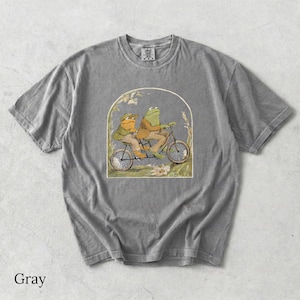 May include: A gray short-sleeve t-shirt featuring a graphic print of two anthropomorphic frogs riding a tandem bicycle. The illustration is framed within an arch and depicts the frogs wearing jackets and hats, set against a grassy background.