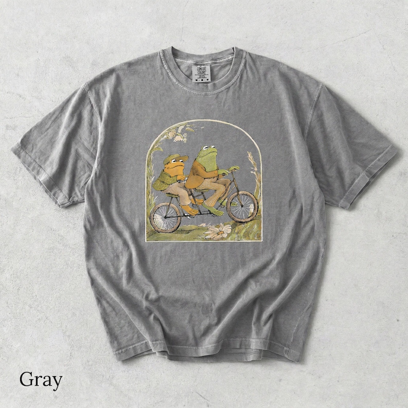 May include: A gray short-sleeve t-shirt featuring a graphic print of two anthropomorphic frogs riding a tandem bicycle. The illustration is framed within an arch and depicts the frogs wearing jackets and hats, set against a grassy background.