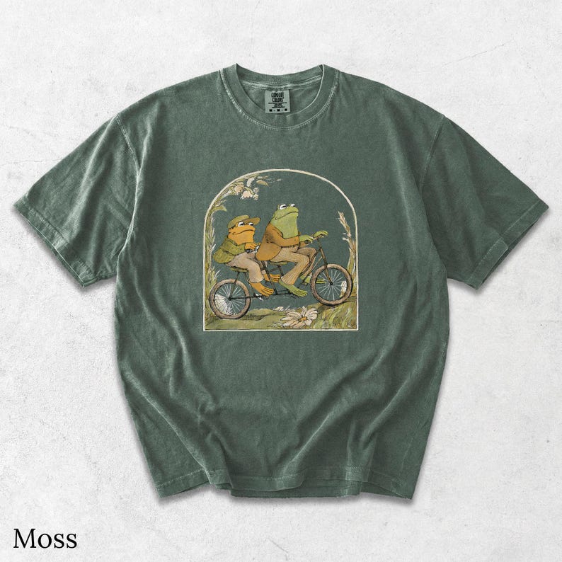 May include: A moss green short-sleeve t-shirt featuring a graphic print of two frogs riding a tandem bicycle. The illustration is framed within an arch and depicts the frogs wearing tan jackets and hats, surrounded by stylised foliage and flowers.