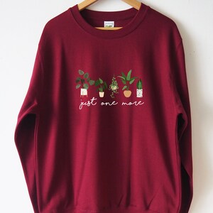 Puede incluir: Una sudadera color burdeos con un gr&aacute;fico blanco de plantas en maceta y el texto "just one more".