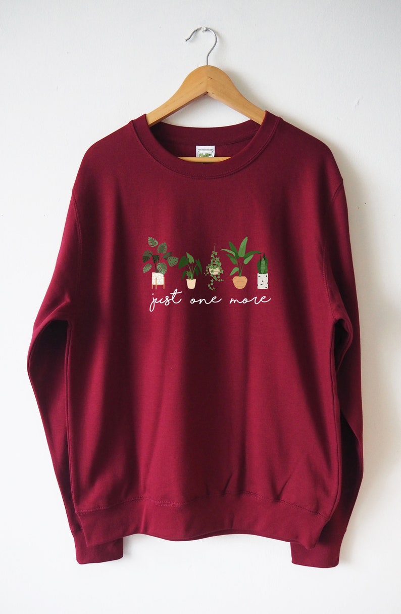 Puede incluir: Una sudadera color burdeos con un gr&aacute;fico blanco de plantas en maceta y el texto "just one more".