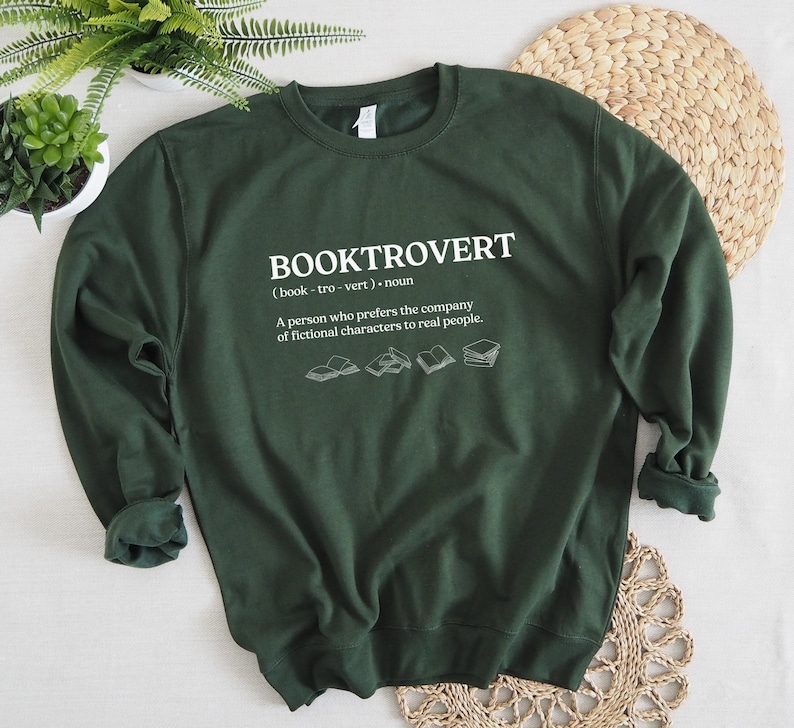 May include: A dark green sweatshirt with the text "BOOKTROVERT (book-tro-vert). noun A person who prefers the company of fictional characters to real people." printed in white. The text is surrounded by five small illustrations of open books.