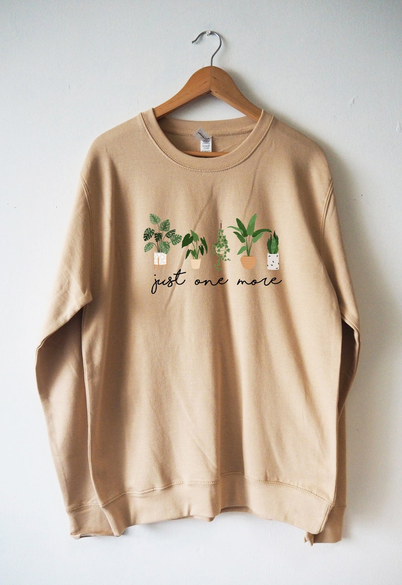 Puede incluir: Una sudadera de color beige con un gr&aacute;fico de plantas en macetas y el texto "just one more".