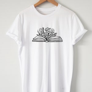 May include: White t-shirt with a black line drawing of an open book with flowers growing out of it.