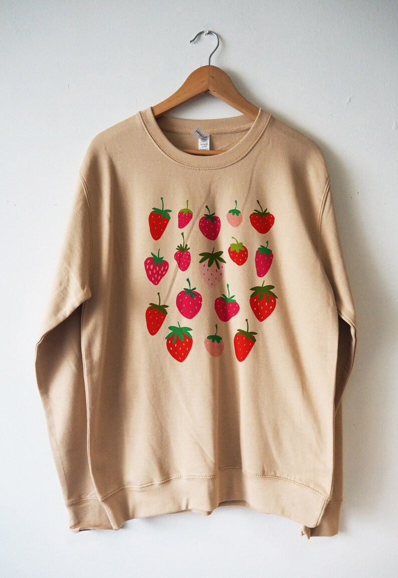 May include: A beige sweatshirt with a pattern of red and green strawberries printed on the front.