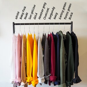 May include: A rack of 12 sweatshirts in various colours, including white, baby pink, nude, sun yellow, gold, heather grey, charcoal, burgundy, navy, earthy green, forest green, and black.