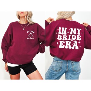 May include: Burgundy sweatshirt with white text that says "Future Mrs. Smith" on the front and "In My Bride Era" on the back. The text is in a retro font with a diamond ring graphic.