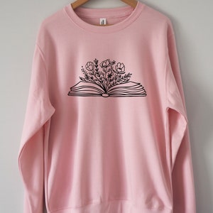 May include: A pink sweatshirt with a black graphic of an open book with flowers growing out of it.