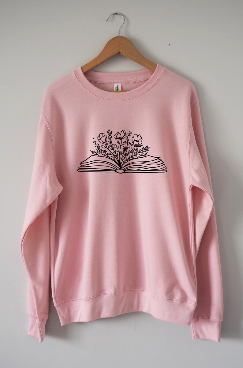 May include: A pink sweatshirt with a black graphic of an open book with flowers growing out of it.