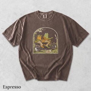 May include: An espresso-colored short-sleeve t-shirt featuring a graphic print of two frogs riding a tandem bicycle. The illustration is framed within an arch and depicts the frogs in a natural setting with plants and flowers.