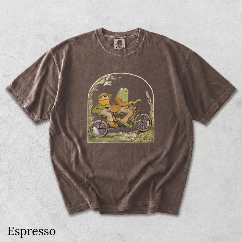 May include: An espresso-colored short-sleeve t-shirt featuring a graphic print of two frogs riding a tandem bicycle. The illustration is framed within an arch and depicts the frogs in a natural setting with plants and flowers.