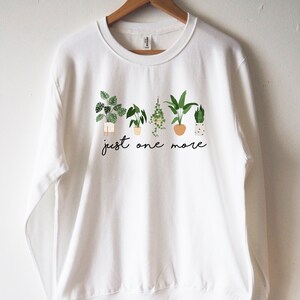 Puede incluir: Sudadera blanca con un gr&aacute;fico de plantas en maceta y el texto "just one more".