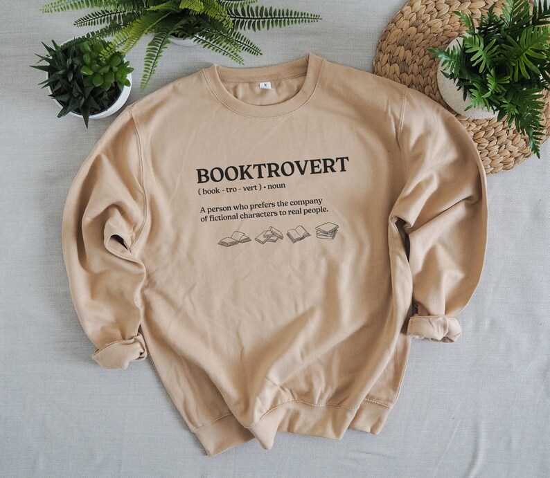 May include: Tan sweatshirt with the text "BOOKTROVERT (book-tro-vert) noun A person who prefers the company of fictional characters to real people." and a graphic of five books.