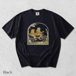 May include: A black short-sleeve t-shirt featuring a graphic print of two anthropomorphic frogs riding a tandem bicycle. The illustration is framed within an arch and depicts the frogs wearing jackets and hats amidst tall grass and flowers.