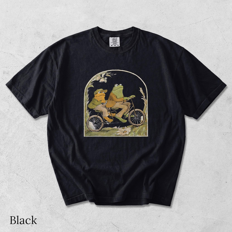 May include: A black short-sleeve t-shirt featuring a graphic print of two anthropomorphic frogs riding a tandem bicycle. The illustration is framed within an arch and depicts the frogs wearing jackets and hats amidst tall grass and flowers.