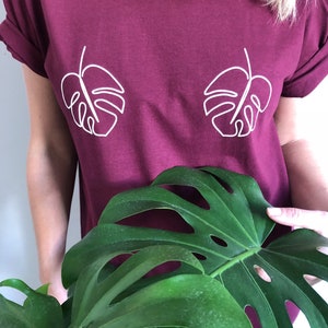 May include: A maroon t-shirt with a white outline of two monstera leaves on the front.