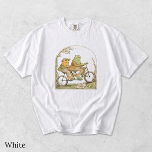 May include: A white short-sleeve t-shirt featuring a central graphic illustration of two anthropomorphic frogs riding a tandem bicycle. The frogs are wearing brown jackets and hats, set within an arched frame with botanical details.
