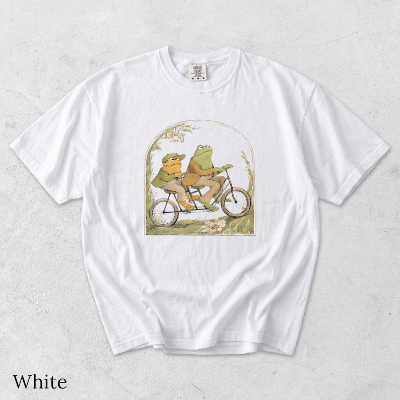 May include: A white short-sleeve t-shirt featuring a central graphic illustration of two anthropomorphic frogs riding a tandem bicycle. The frogs are wearing brown jackets and hats, set within an arched frame with botanical details.