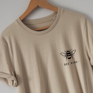 Może przedstawiać: Beżowa koszulka z czarnym nadrukiem pszczoły i tekstem "Bee Kind."