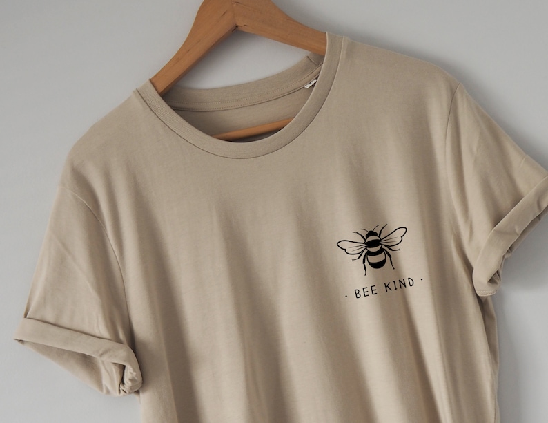 Może przedstawiać: Beżowa koszulka z czarnym nadrukiem pszczoły i tekstem "Bee Kind."