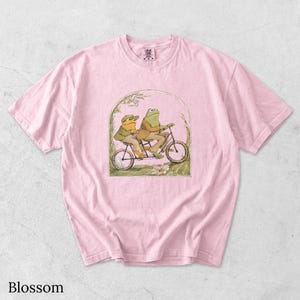 May include: A light pink short-sleeve t-shirt featuring a graphic illustration of two frogs riding a tandem bicycle. The frogs are wearing jackets and hats, framed within an arched border with floral details. The shirt has a crew neckline.
