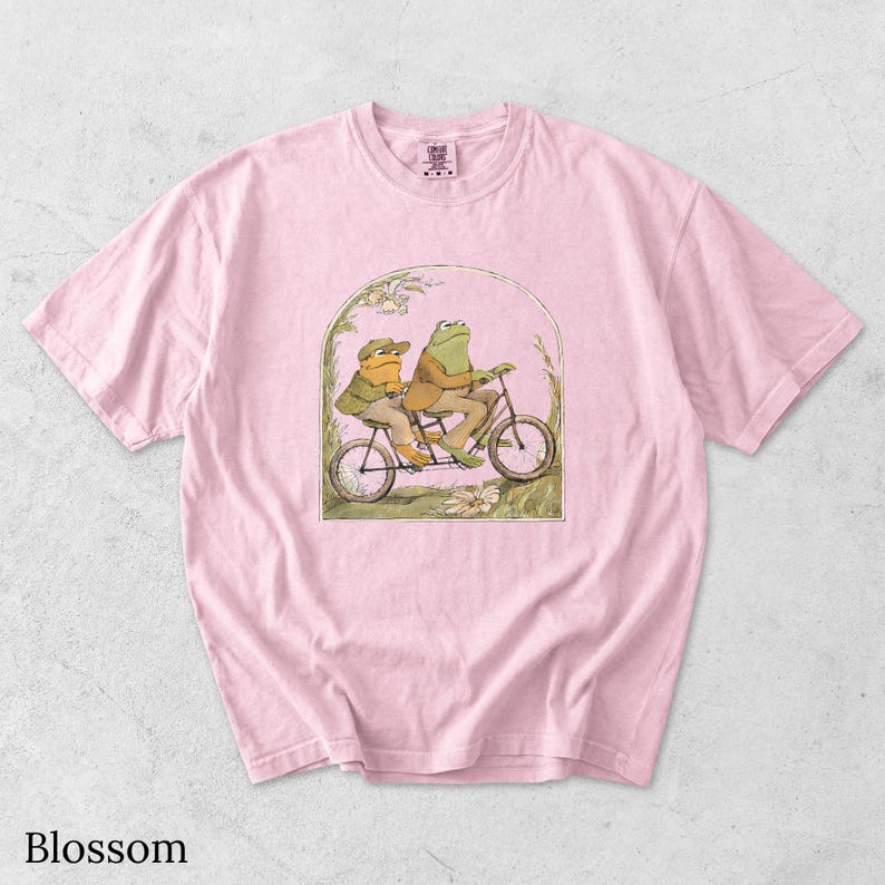 May include: A light pink short-sleeve t-shirt featuring a graphic illustration of two frogs riding a tandem bicycle. The frogs are wearing jackets and hats, framed within an arched border with floral details. The shirt has a crew neckline.
