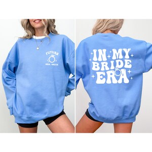 May include: A light blue sweatshirt with the text "Future Mrs. Smith" and a ring graphic on the front. The back of the sweatshirt has the text "In My Bride Era" in a retro font with a ring graphic.