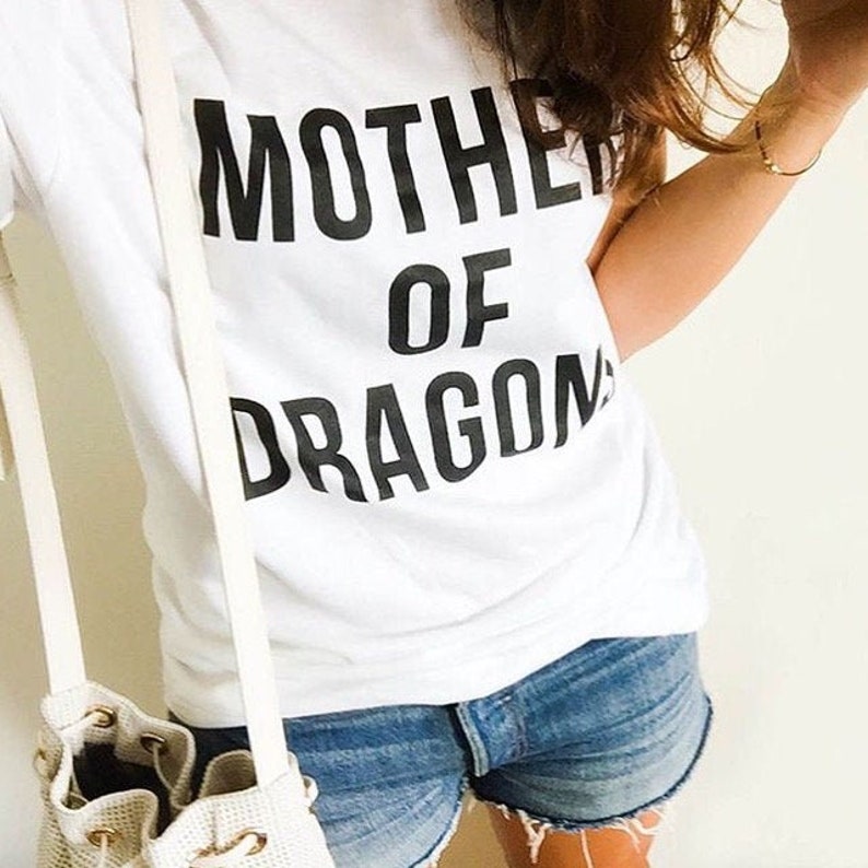 May include: White t-shirt with the text "MOTHER OF DRAGON" in bold black letters. The shirt is paired with denim shorts. A white purse with a strap is visible on the left side.