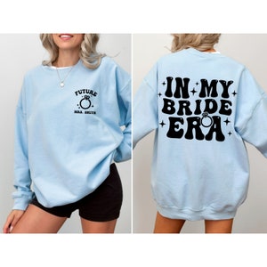 May include: Light blue sweatshirt with black text that says "In My Bride Era" with a diamond ring graphic. The front of the sweatshirt has black text that says "Future Mrs. Smith" with a diamond ring graphic.