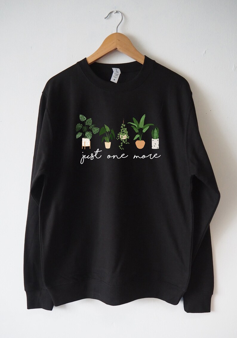 Puede incluir: Sudadera negra con un gr&aacute;fico de plantas en maceta y el texto "just one more".