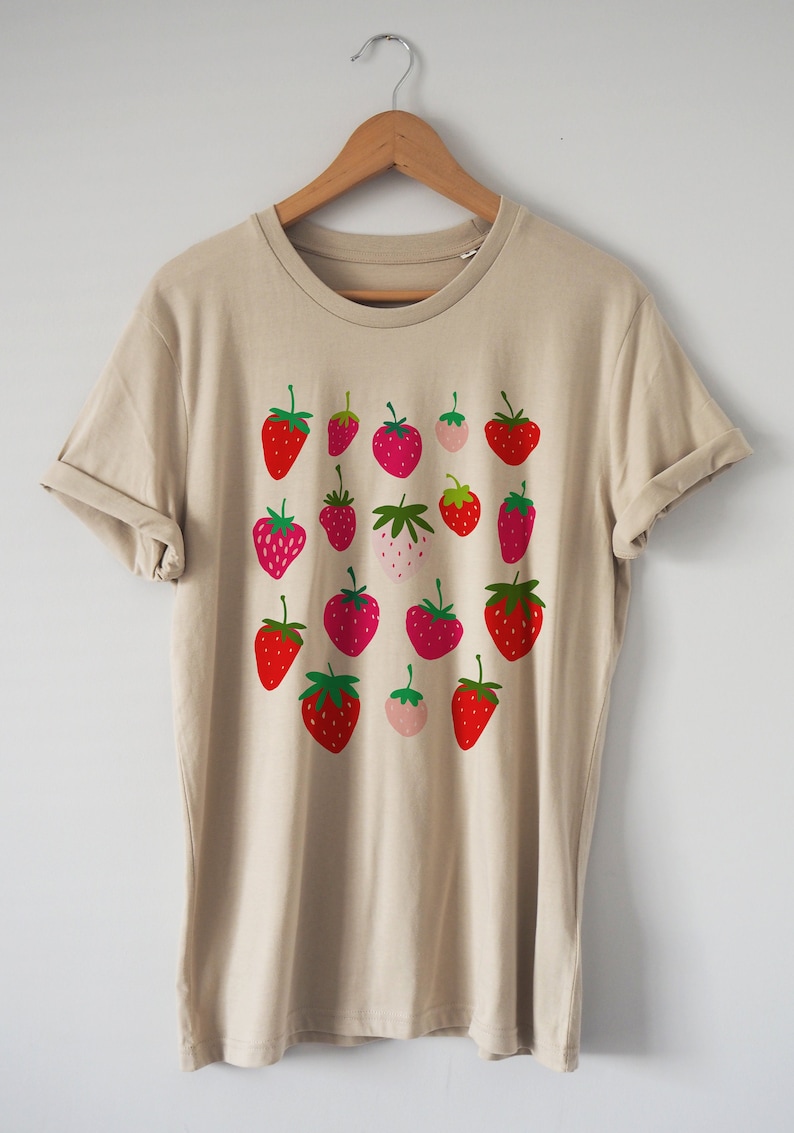 Puede incluir: Una camiseta beige con un estampado gr&aacute;fico de fresas rojas, rosas y verdes.